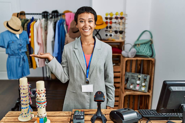 Junge hispanische Frau mit kurzen Haaren, die als Managerin in einer Boutique arbeitet, lächelt fröhlich und zeigt mit der Handfläche in die Kamera.  - Foto, Bild
