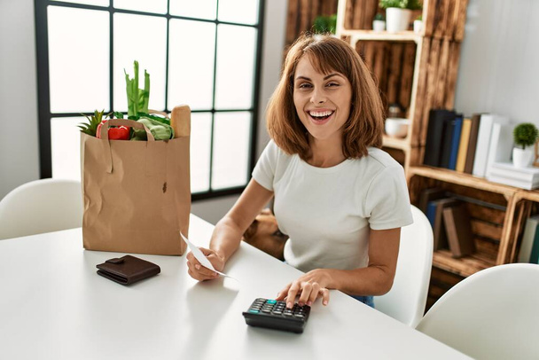 Giovane donna caucasica sorridente fiducioso facendo contabilità di acquisto di generi alimentari a casa - Foto, immagini