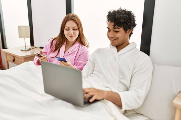 Mladý pár usměvavý šťastný pomocí notebooku, smartphone a kreditní karty v ložnici. - Fotografie, Obrázek