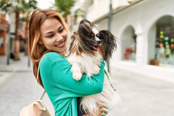 Νεαρό καυκάσιο κορίτσι χαμογελά χαρούμενος κρατώντας το σκυλί στην πόλη. - Φωτογραφία, εικόνα
