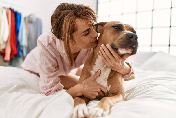 Νεαρή καυκάσια γυναίκα αγκαλιάζει και φιλάει το σκύλο ξαπλωμένη στο κρεβάτι στο υπνοδωμάτιο - Φωτογραφία, εικόνα