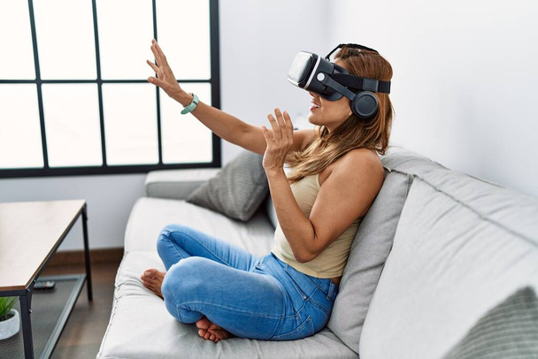 Молодая латинская женщина играет в видеоигры, используя очки виртуальной реальности дома - Фото, изображение