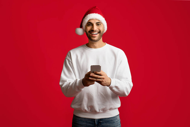 App mobile. Allegro ragazzo arabo che indossa il cappello di Babbo Natale con smartphone nelle mani - Foto, immagini