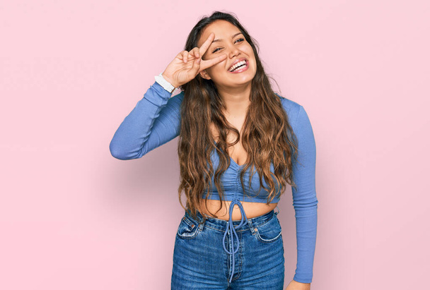 Молодая латиноамериканка в повседневной одежде делает символ мира с пальцами на лице, улыбаясь радостно показывая победу  - Фото, изображение