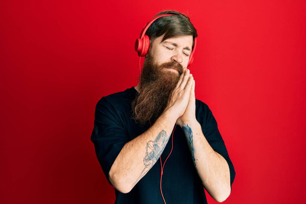 Homme rousse à longue barbe écoutant de la musique à l'aide d'écouteurs endormis fatigué rêvant et posant les mains ensemble tout en souriant les yeux fermés.  - Photo, image