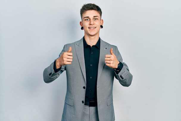 Молодой кавказский мальчик с расширившимися ушами, одетый в знак успеха деловой куртки, делает позитивный жест рукой, улыбается и счастлив. веселое выражение лица и жест победителя.  - Фото, изображение