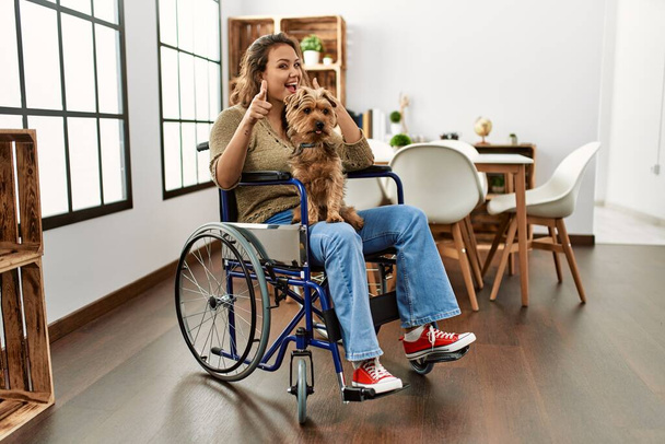 Young hispanic girl sitting on wheelchair at home looking confident at the camera with smile with crossed arms and hand raised on chin. thinking positive.  - Foto, Imagem