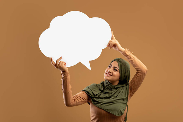 Σχόλιο. Ενθουσιασμένη αραβική κυρία που έχει ιδέα ή σκέψη, κρατώντας κενό σύννεφο φυσαλίδων ομιλίας πάνω από το κεφάλι της, κενό διάστημα - Φωτογραφία, εικόνα