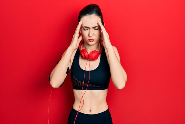 Young hispanic girl wearing gym clothes and using headphones with hand on head for pain in head because stress. suffering migraine.  - Zdjęcie, obraz