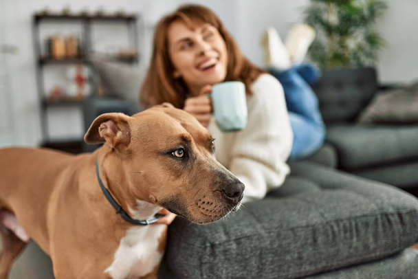 Joven mujer caucásica bebiendo café tumbado en el sofá con perro en casa - Foto, imagen