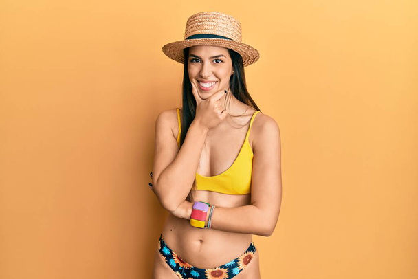 Młoda Latynoska kobieta ubrana w bikini i letni kapelusz, wyglądająca pewnie na aparat uśmiechnięta ze skrzyżowanymi ramionami i podniesioną ręką na podbródku. pozytywne myślenie.  - Zdjęcie, obraz