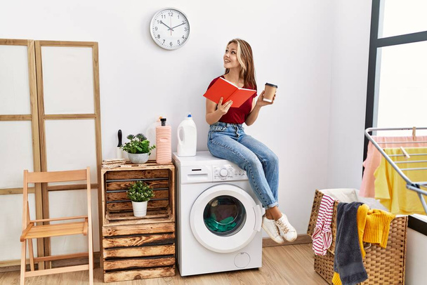 Young caucasian girl drinking coffee and reading book waiting for laundry sitting on whasing machine at home. - Photo, Image