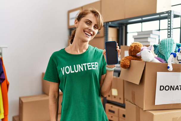 Joven mujer voluntaria caucásica sonriendo feliz mostrando aplicación de voluntariado en el centro de caridad. - Foto, Imagen