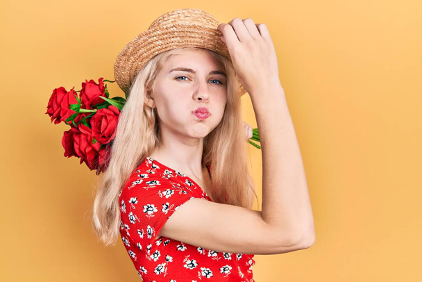 Beautiful caucasian woman with blond hair holding bouquet of red roses puffing cheeks with funny face. mouth inflated with air, catching air.  - Valokuva, kuva
