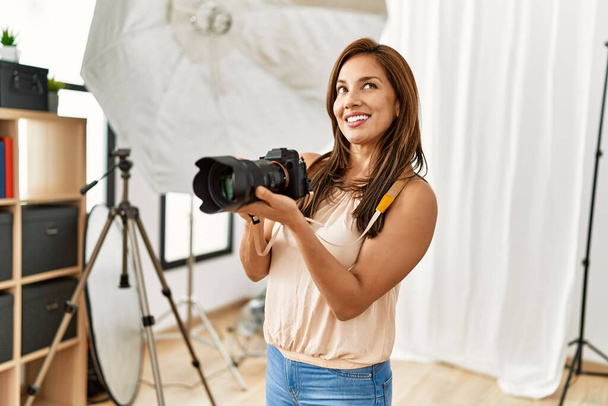 Joven fotógrafa hispana sonriendo feliz usando la cámara en el estudio fotográfico. - Foto, imagen