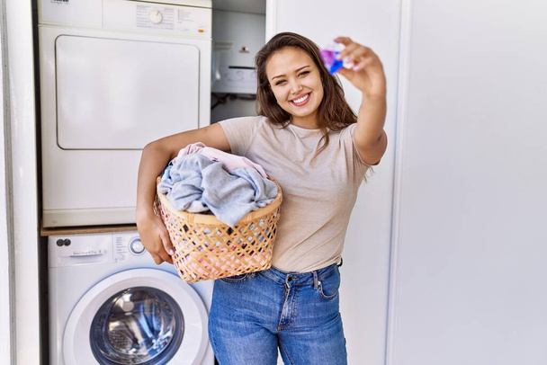 Chica hispana joven haciendo cesta de ropa con ropa y bolsa de detergente en casa - Foto, Imagen