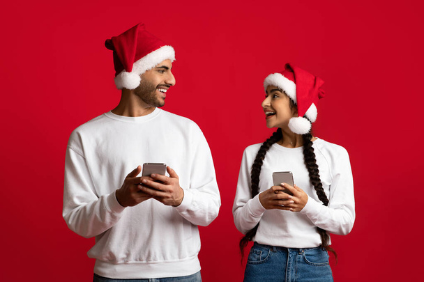 Gran aplicación. Emocionados cónyuges árabes en Santa Sombreros Sosteniendo Smartphones - Foto, imagen