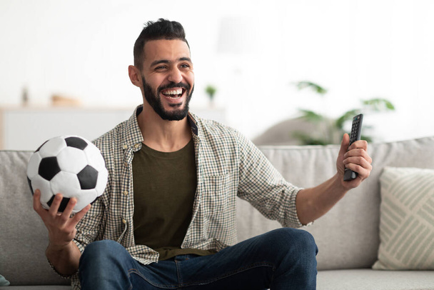 Happy young Arab man watching soccer game on television, holding ball and controller at home - Фото, изображение