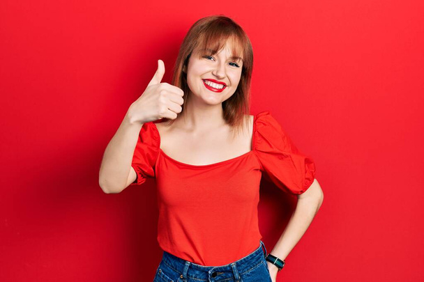 Zrzavá mladá žena v neformálním červeném tričku dělá šťastné gesto rukou nahoru. schválení výrazu při pohledu na fotoaparát, který vykazuje úspěch.  - Fotografie, Obrázek