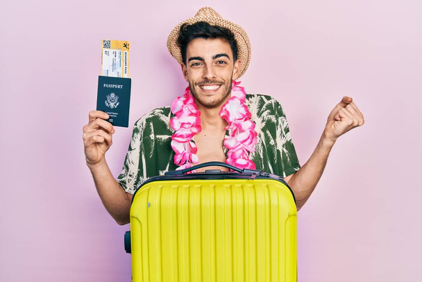 Joven hombre hispano con estilo veraniego y lei hawaiano con pasaporte y tarjeta de embarque gritando orgulloso, celebrando la victoria y el éxito muy emocionado con el brazo levantado  - Foto, Imagen