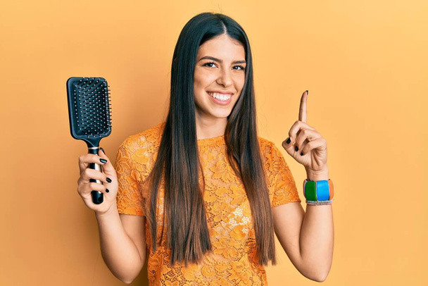 Joven mujer hispana sosteniendo un cepillo de pelo sonriendo con una idea o pregunta apuntando con el dedo con la cara feliz, número uno  - Foto, imagen