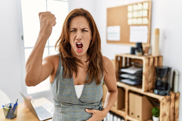 Hispanische Frau mittleren Alters im Büro wütend und wütend die Faust erhoben frustriert und wütend, während sie vor Wut schrie. Wut und aggressives Konzept.  - Foto, Bild