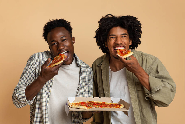 おいしいお菓子。ボックスからイタリアのピザを食べる2人の空腹の黒人男性 - 写真・画像
