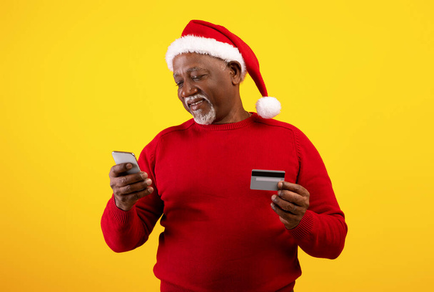 携帯電話とクレジットカードを身に着けている陽気なシニア黒人男性サンタ帽子、クリスマスプレゼントをオンラインで購入 - 写真・画像