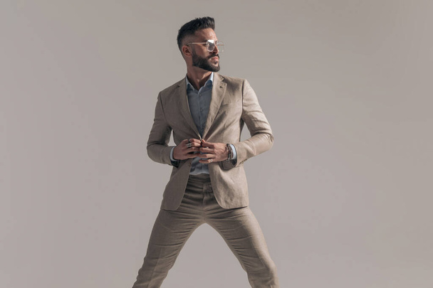 σέξι επιχειρηματίας μόδας κλείνοντας το σακάκι του, κοιτάζοντας μακριά και ποζάροντας σε γκρι φόντο - Φωτογραφία, εικόνα
