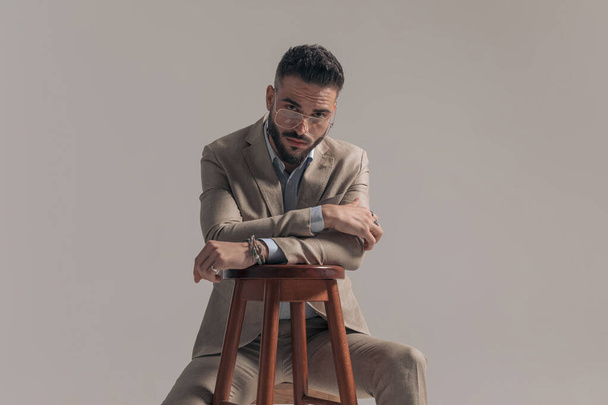young fashion businessman looking over his glasses and leaning on a chair against gray background - Fotó, kép