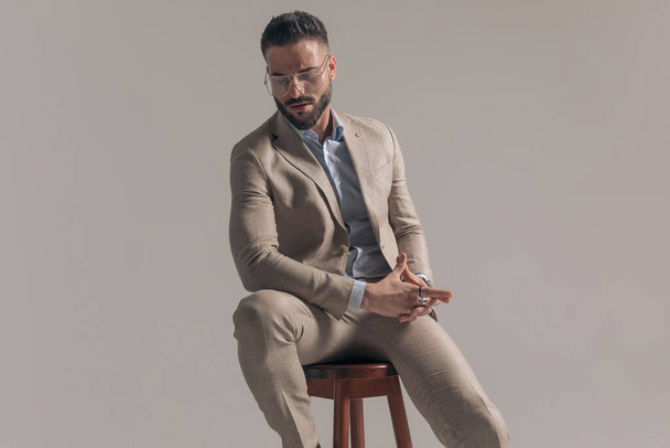 dramatic macho businessman posing on a chair and rubbing his hands on gray background - Foto, Imagem