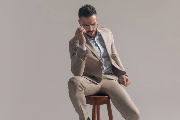 junger Geschäftsmann posiert auf einem Stuhl, ist dramatisch und trägt eine Brille auf grauem Hintergrund - Foto, Bild