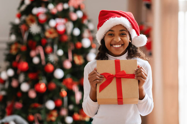 Μαύρο κορίτσι σε Σάντα καπέλο κρατώντας χριστουγεννιάτικο δώρο στο σπίτι - Φωτογραφία, εικόνα