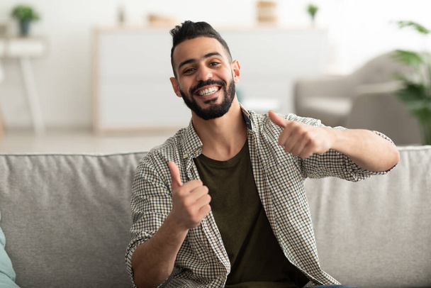 Веселый молодой араб, показывающий пальцами вверх жест обеими руками и улыбающийся в камеру, сидящий дома на диване - Фото, изображение