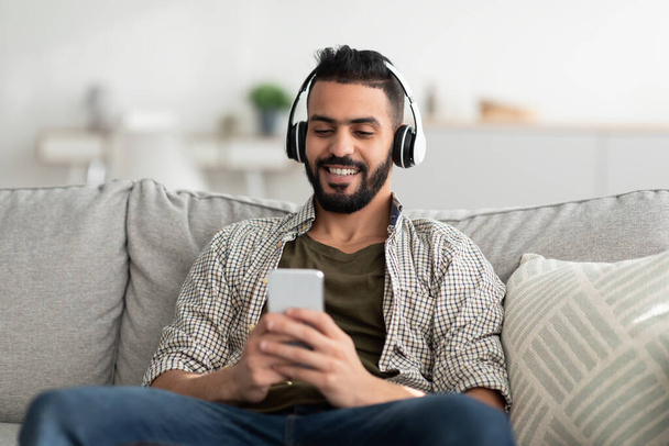 Positive Arab guy in headset listening to music, radio or audio book on mobile phone, choosing popular playlist at home - Foto, immagini