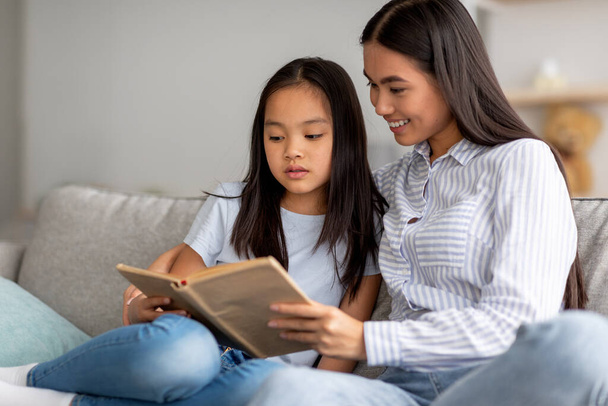 Οικογενειακές ασχολίες. Ευτυχισμένη Ασιάτισσα μητέρα και η κόρη της διαβάζουν μαζί βιβλίο, περνούν χρόνο στο σπίτι, ελεύθερο χώρο - Φωτογραφία, εικόνα