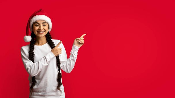 Reklama świąteczna. Szczęśliwy młody Arab kobieta w Santa kapelusz wskazując obok - Zdjęcie, obraz