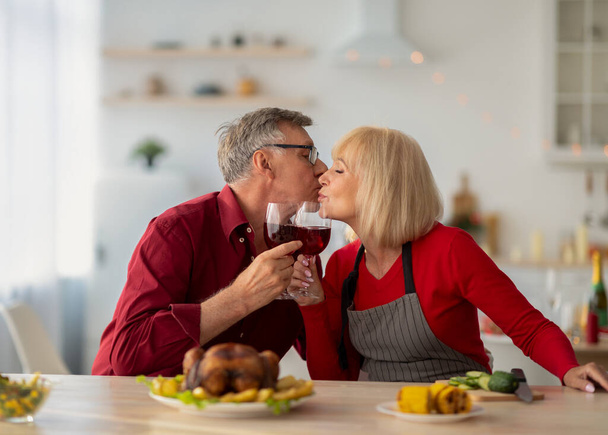 Ласковый старший мужчина и его жена целуются и пьют вино во время приготовления праздничной еды на Рождество или День благодарения - Фото, изображение
