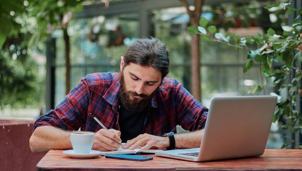 Ernstige hipster kerel neemt notities in zijn notebook, terwijl het lezen van zijn laptop, terwijl het hebben van een koffie in een cafe. - Foto, afbeelding