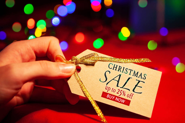 A woman's hand holds a label with text - Christmas sale up to 25 off, buy now. On a red background, with a multi-colored gerland. - Fotografie, Obrázek
