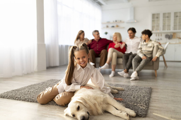 Entzückendes Mädchen sitzt auf weichem Teppich mit Golden Retriever Hund, ihre Verwandten entspannen auf dem Sofa im Wohnzimmer - Foto, Bild