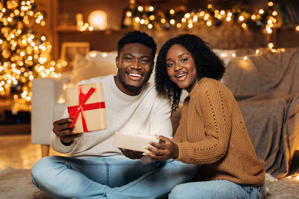 Fröhliches junges afroamerikanisches Paar öffnet gemeinsam Weihnachtsgeschenk im geschmückten Wohnzimmer - Foto, Bild