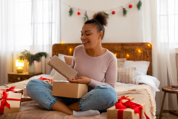 Meglepett afro-amerikai nő kipakol karácsonyi ajándékdobozokat és kuplungot tart, az ágyon ül és mosolyog. - Fotó, kép