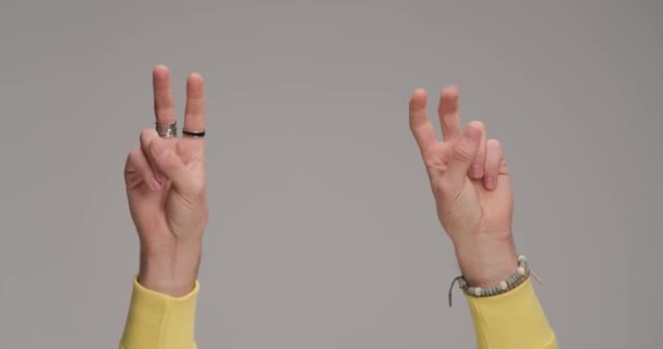 las manos del hombre sosteniendo los dedos en el aire y haciendo un gesto de paz frente al fondo gris en el estudio - Imágenes, Vídeo