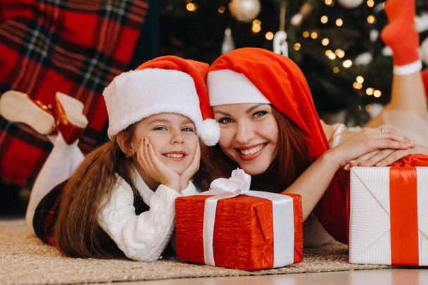 A happy little girl and her mom in Santa Claus hats are smiling with gifts in their hands. - Foto, immagini