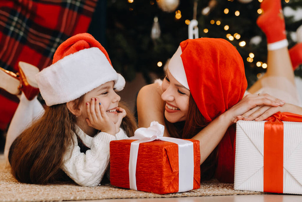 Szczęśliwa dziewczynka i jej mama w kapeluszach Świętego Mikołaja uśmiechają się z prezentami w rękach. - Zdjęcie, obraz