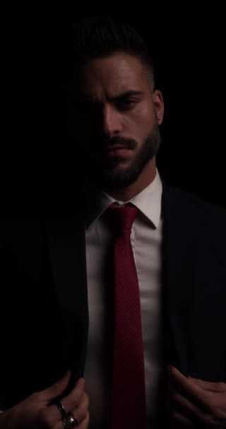 сексуальний молодий бізнесмен з бородою дивиться в бік, влаштовує і закриває костюм, торкається долонь і позує перед чорним тлом в студії
 - Кадри, відео
