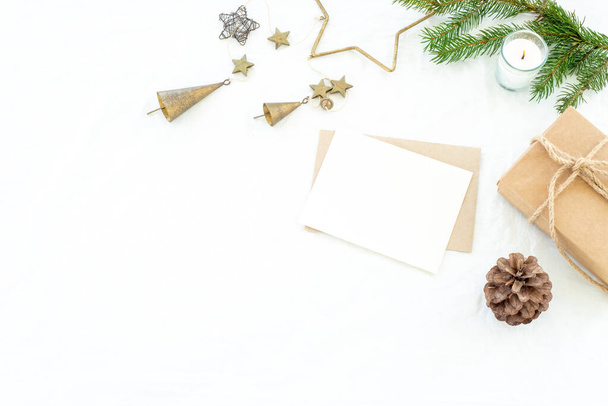 Різдвяні прикраси композиції. Різдвяні вітальні листівки макет сцени. Порожня вітальна листівка та конверт, подарункова різдвяна коробка зі стрічкою, ялинова гілка, сосновий конус, металеві прикраси та запалена свічка. Білий лляний фон. Простір для копіювання, вид зверху, плоский
 - Фото, зображення