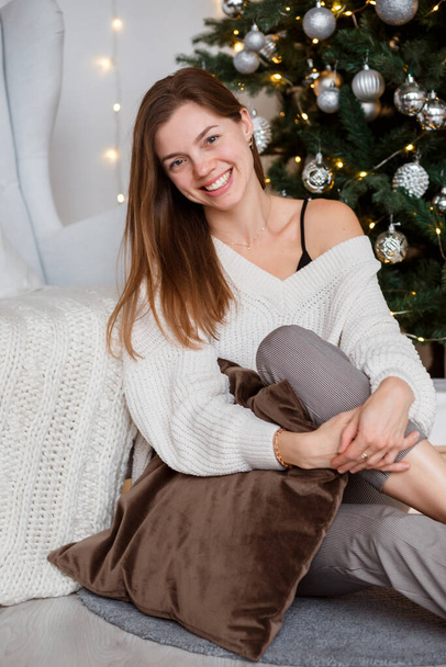 Portrait of cheerful smiling woman in warm knitted sweater sitting next to Christmas tree.  - Valokuva, kuva
