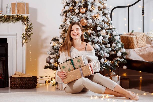 Cheerful woman sitting next next to Christmas tree with gift boxes, celebrating Christmas at home.  - Фото, зображення
